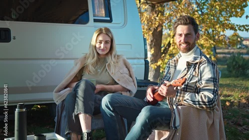 Young beautiful couple enjoying nice sunny weather in nature playing favorite music on guitar. Attractive woman with her beloved man smiling and looking at camera. Modern camping bus on background. photo