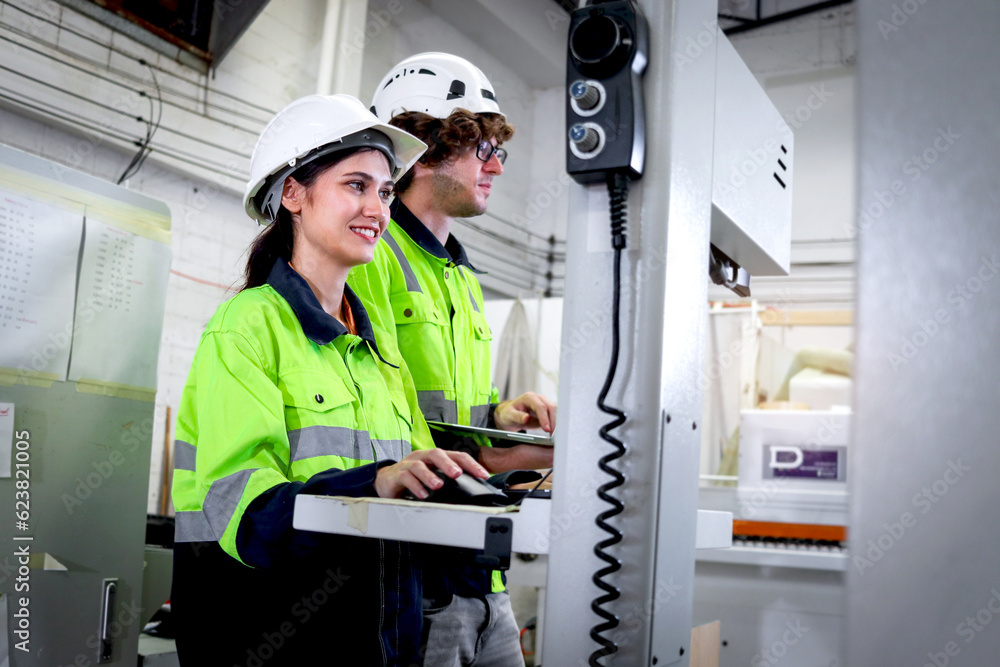 Happy smiling industrial woman and man workers wear helmet and safety vest work together with computer controller, operate automatic machinery robot at furniture factory, employees of CNC woodworking. Happy smiling industrial woman and man workers wear helmet and safety vest work together with computer controller, operate automatic machinery robot at furniture factory, employees of CNC woodworking.