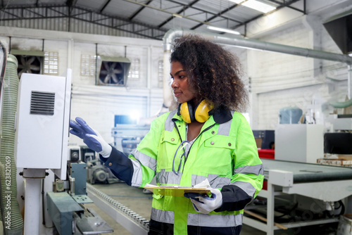 African industrial woman worker with curly hair wears safety vest, writes on file folder document, female engineer check and maintenance machinery at CNC woodworking manufacturing furniture factory.