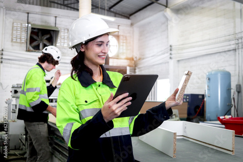 Happy industrial woman worker with helmet and safety vest holding tablet and inspecting wood raw material, coworker working with automatic robot machinery as background, employees of CNC woodworking.