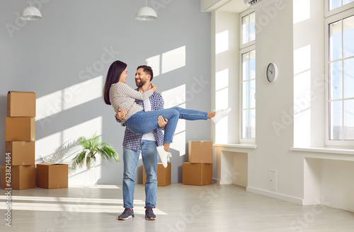 Young family couple having fun on moving day in new house or apartment. Happy husband carrying wife. Joyful man standing in empty unfurnished living room with boxes and holding his woman in his arms