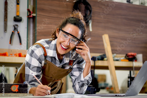 Happy smiling beautiful carpenter woman wearing safety glasses goggles and apron taking to customer by using mobile phone, female craft worker with wooden furniture background in carpentry workshop.