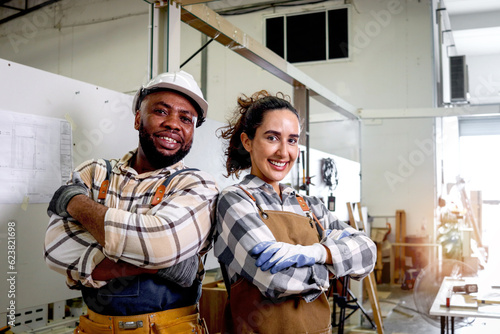 Portrait of two happy smiling carpenter man and woman wearing apron standing with arms crossed in woodworking furniture workshop, beautiful carpentry woman with her coworker friend African craftsman