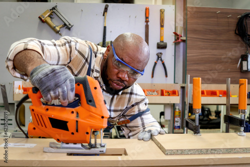 Middle-aged African carpenter wears safety glasses goggles and apron, using carpentry tools with wood plank to make wooden furniture in woodworking workshop, worker craftsman works at workshop.