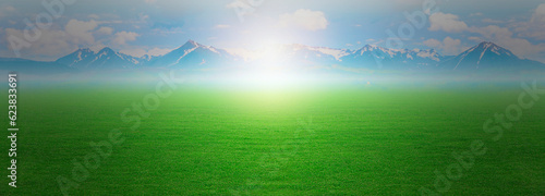 Landscape view of green grass and fog smoke with clouds and bright sun blue sky and mountain range in the background  summer spring meadow