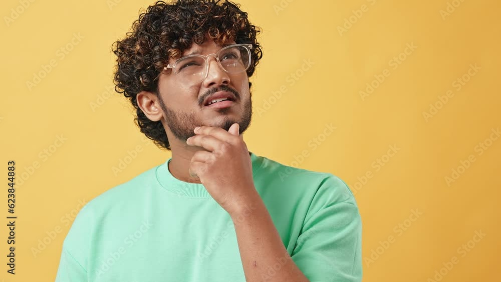 Portrait of a handsome curly-haired young man in a turquoise t-shirt and glasses standing on a yellow background and thoughtfully scratching his chin.The emotion of doubt, reflection and confusion. Portrait of a handsome curly-haired young man in a turquoise t-shirt and glasses standing on a yellow background and thoughtfully scratching his chin.The emotion of doubt, reflection and confusion.