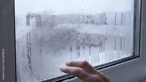 Female Hand Wipes the Dew, Condensate from a Wet, Misted Window. Fingers slide on wet glass. Drops of the condensate flow down. View from the window on the roofs of the houses. Gloomy rainy day. 4K.