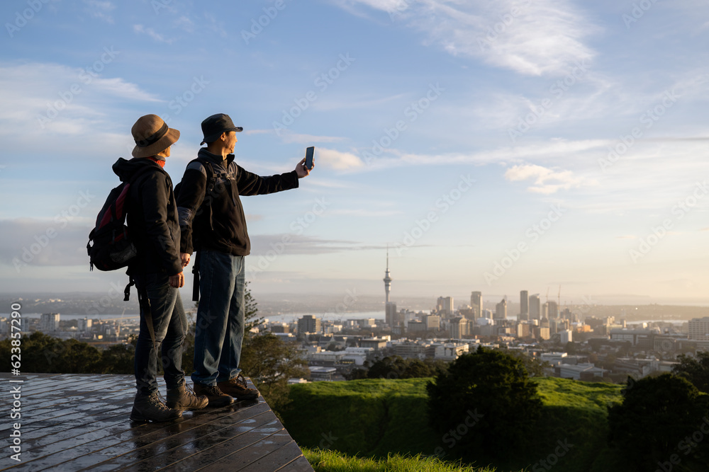 Couple taking photo with smartphone on Mt Eden summit at sunrise. Skytower in the background. Selective focus on people in foreground. Auckland.