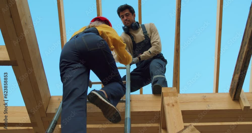 male contractor helping to female to climb on the roof of wooden building house on stepladder, rear view on female climbing to height. unfinished building site. male contractor helping to female to climb on the roof of wooden building house on stepladder, rear view on female climbing to height. unfinished building site.