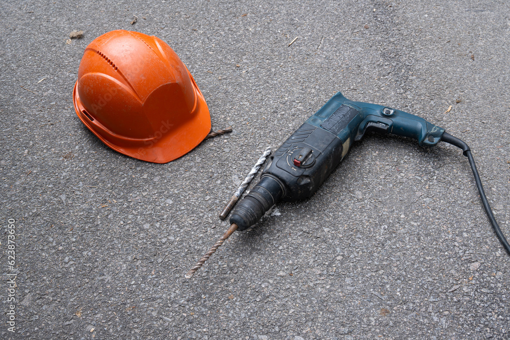 a hammer drill lies on the ground next to a protective construction helmet