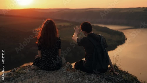 Couple man and woman looking at river view at sunset, romantic vacation during travel. Couple watching sunset, talking on cliff edge above river in nature, back view.
