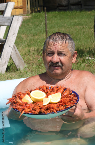 Joyful mustachioed man sits in the pool. He holds a large dish with boiled crayfish in his hands. Crayfish are decorated with lemon slices. Close-up.