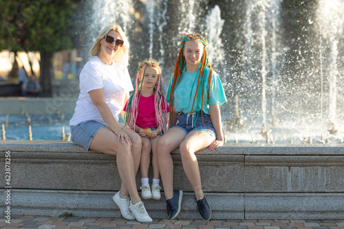 Mother with two happy girls sisters in a bright summer t-shirt with colored African braids in the hair have fun by a city fountain. Happy family have a rest near a fountain during a heat wave.
