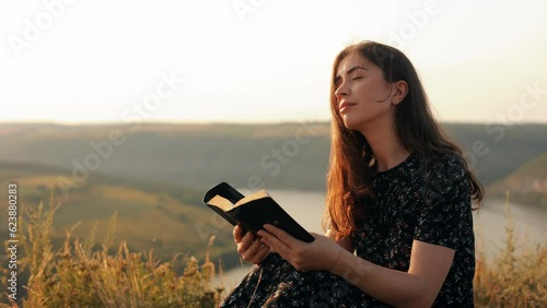 A long-haired girl reads a book in the sun. A young woman reads the Bible outdoors. A woman holds a Bible in her hands and studies the word of God on top of a mountain. 