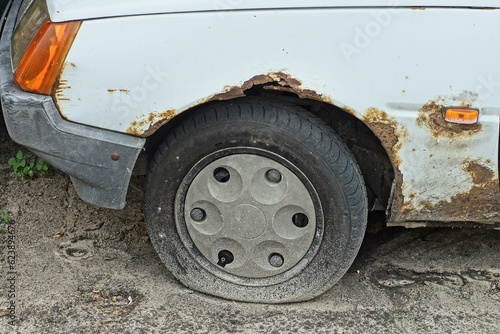 pierced with a sharp object on the road and a flat rubber black wheel of an old rusty iron white car stands on the ground on the street during the day © FEDOR
