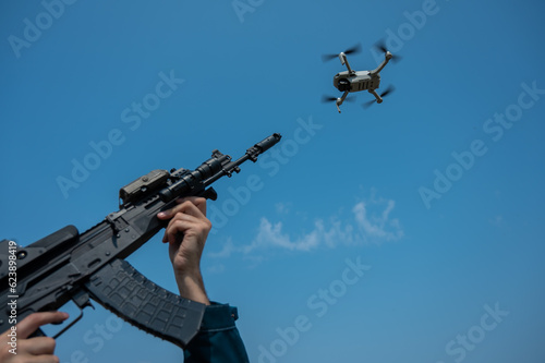 A man aims to shoot a rifle at a flying drone against a blue sky. 