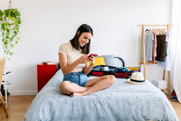 Young pretty traveler woman using smartphone to book a hotel while preparing travel suitcase before going on summer vacation. Online booking concept.