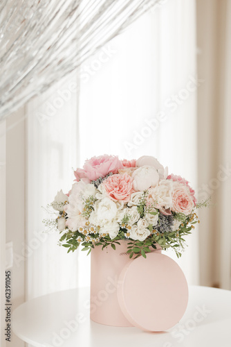 An elegant bouquet of fresh roses, peonies, and daisies in a pink box, beautifully arranged and placed by a light-filled window. © Kotkoa