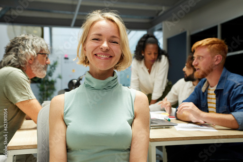 Portrait of an office cheerful Nordic mature woman and looking at camera. Empowered female leader. In the background there is a group of employees kipping at work. It is an indoor multiracial scene. photo