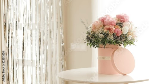 An elegant bouquet of fresh roses, peonies, and daisies in a pink box, beautifully arranged and placed by a light-filled window.