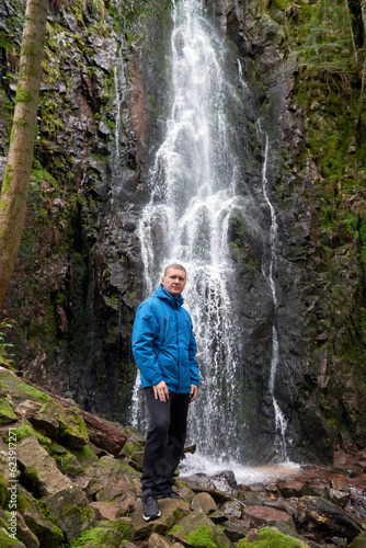 Tourist attraction of Germany - falls of Burgbach Waterfall near Schapbach, Black Forest, Baden-Wurttemberg, Germany. Man hiker in blue jacket standing on stone and looks at flow of falling water