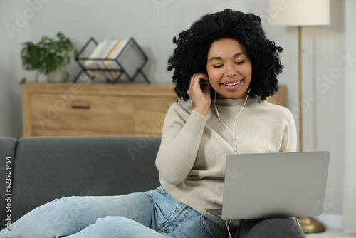 Happy young woman with earphones using laptop on sofa indoors