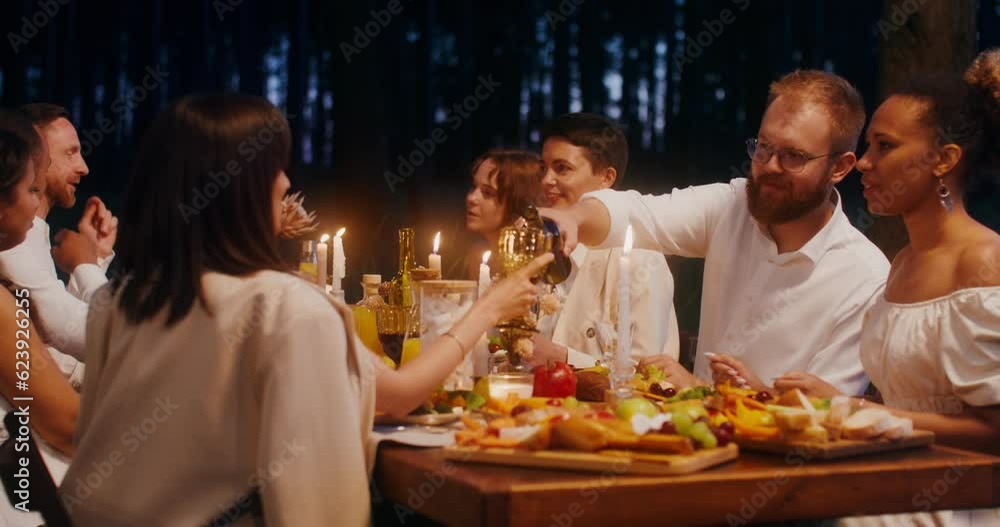 Guests smile and talk while sitting at a beautifully set festive table outdoors. A man pours wine into a glass Guests smile and talk while sitting at a beautifully set festive table outdoors. A man pours wine into a glass