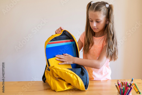 Blonde Elementary School Girl With Pigtails Puts Office Supplies Into A Backpack. Getting ready for school. Back to school. Self-assembly Of A School Backpack. First day of class