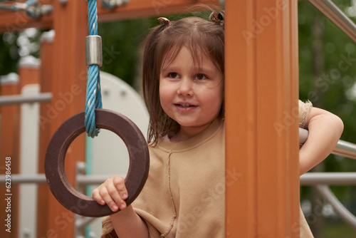 A girl is playing on an outdoor playground. She has blown up the stairs and is holding on to a suspended sports ring.