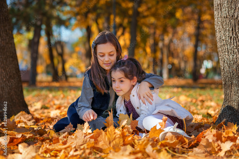 two girls are sitting in a glade of yellow maple leaves in an autumn city park, children are playing and enjoying, picking leaves near a tree, beautiful nature, bright sunny day