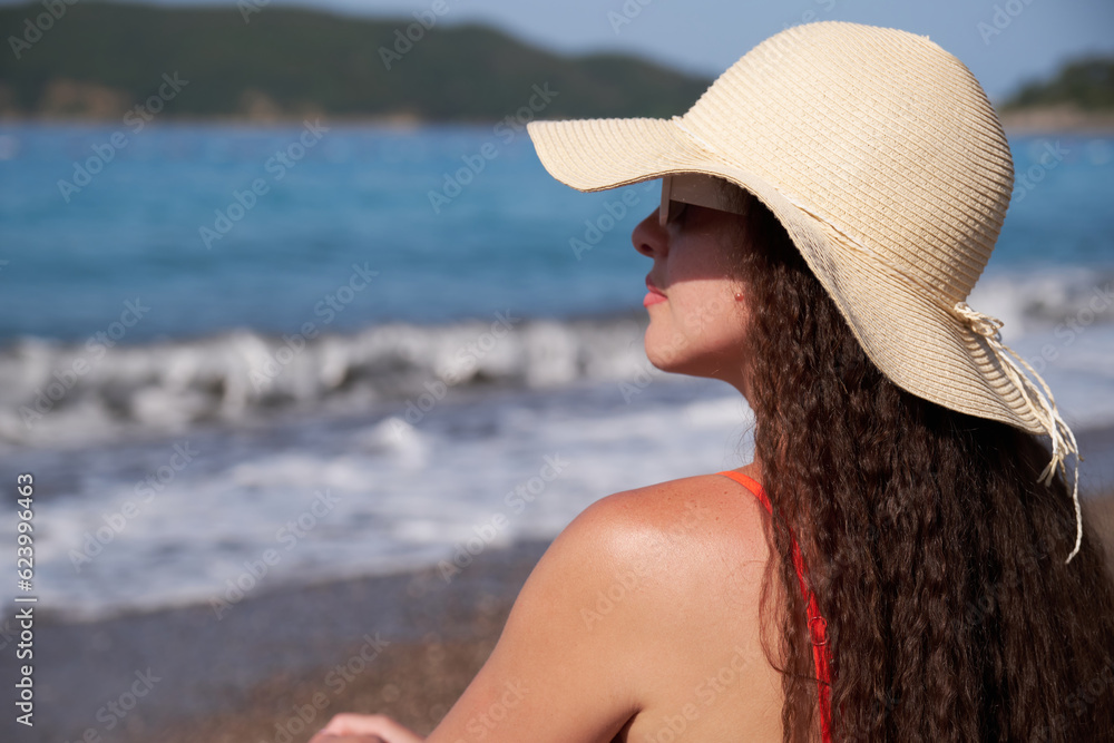 A beautiful woman in a hat sits on a pebble beach near the sea.