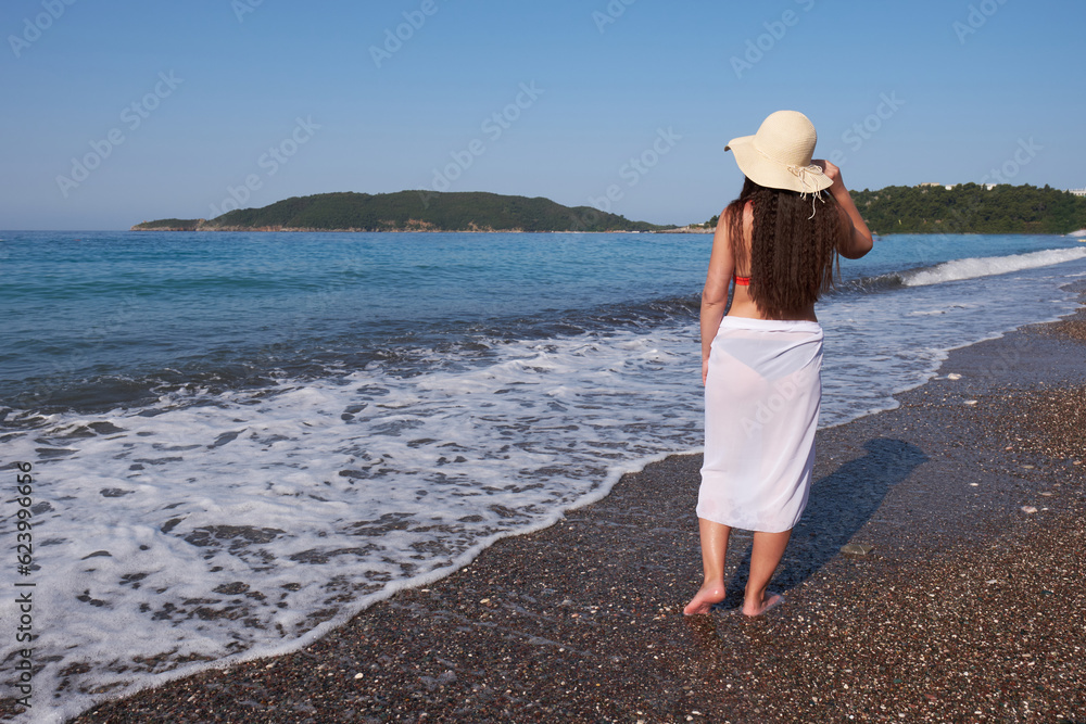Young brunette woman with long hair in a white pareo and hat walks along the beach.