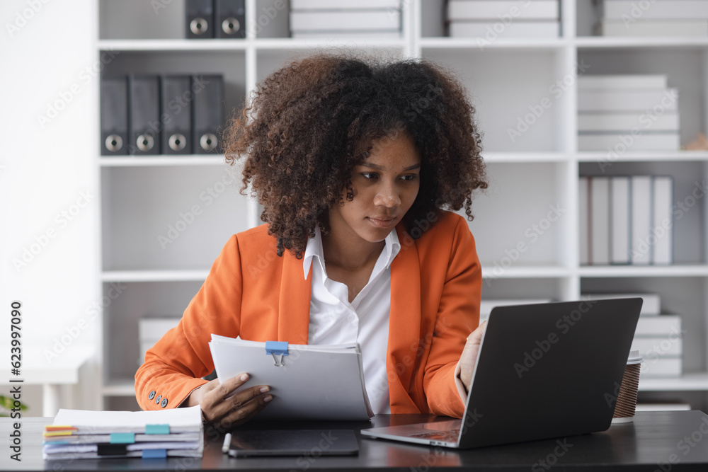 Young African American businesswoman holding a document paper and laptop computer at desk in office, Advice on financial business concept. Young African American businesswoman holding a document paper and laptop computer at desk in office, Advice on financial business concept.