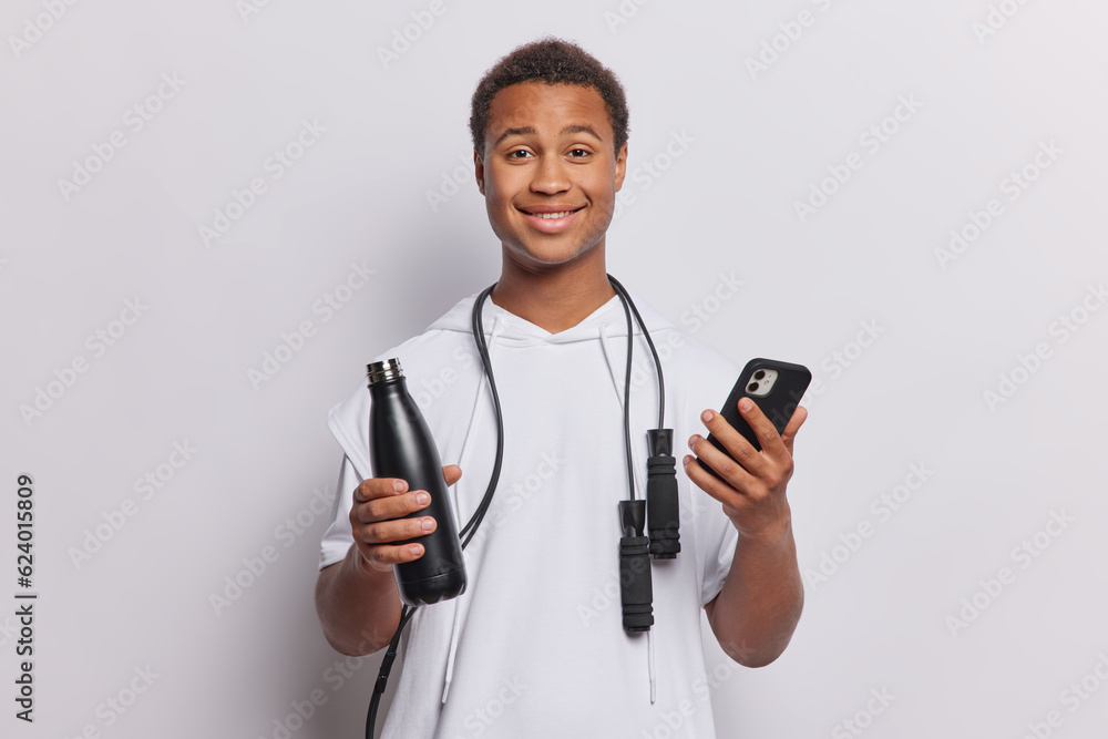Cheerful African guy poses with smartphone and bottle of water uses jumping rope for exercising stays in good physical shape smiles pleasantly dressed in t shirt isolated over white background Cheerful African guy poses with smartphone and bottle of water uses jumping rope for exercising stays in good physical shape smiles pleasantly dressed in t shirt isolated over white background