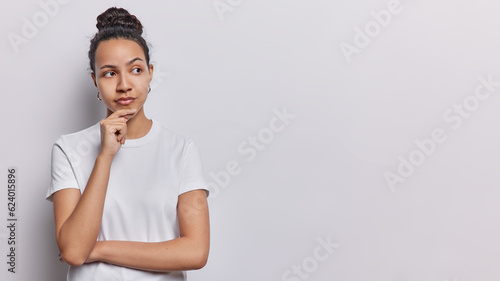 Thoughtful Latin girl keeps hand on chin concentrated aside has dark hair gathered in bun dressed in casual t shirt isolated over white background with copy space for your advertising content photo