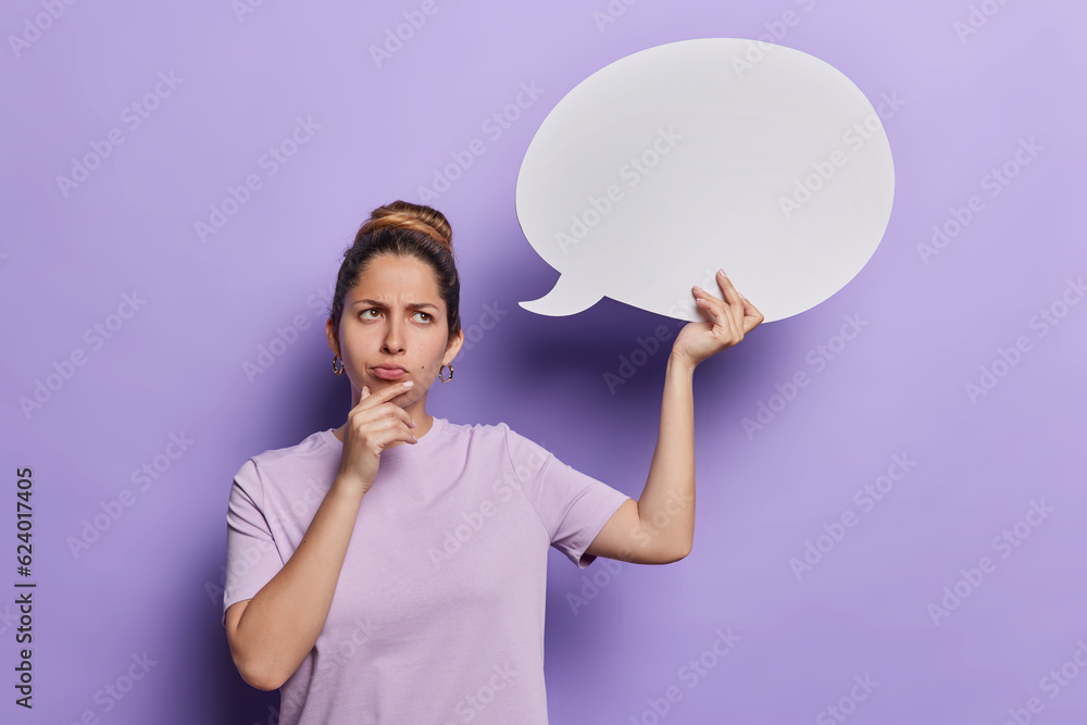 Photo of displeased young European woman holds chin frowns face being deep in thoughts holds communication bubble for your promotional text dressed in casual t shirt isolated over purrple background Photo of displeased young European woman holds chin frowns face being deep in thoughts holds communication bubble for your promotional text dressed in casual t shirt isolated over purrple background