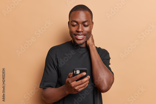 Pleased dark skinned man keeps hand on neck smiles pleasantly holds mobile phone watching video using entertaining mobile application dressed in casual black t shirt isolated over brown background. © Wayhome Studio