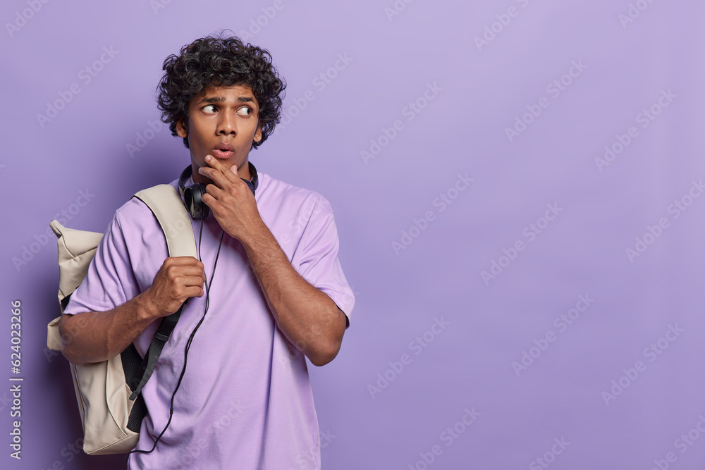 Indoor portrait of young dreamy hindu student trying to solve problem wearing casual purple tshirt with big white bag isolated on purple background on left side with blank space for your advertisement Indoor portrait of young dreamy hindu student trying to solve problem wearing casual purple tshirt with big white bag isolated on purple background on left side with blank space for your advertisement