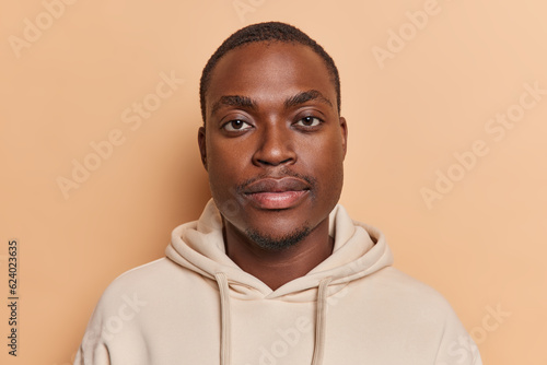 Portrait of handsome dark skinned bearded man concentrated directly at camera has calm expression dressed in casual sweatshirt poses against brown background. Ethnic people and lifestyle concept