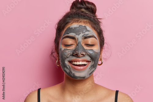 Skincare and wellness concept. Horizontal indoor close up of young pretty pleased woman smilling broadly and happily with closed eyes applying natural beauty clay mask to make her wrinkless less
