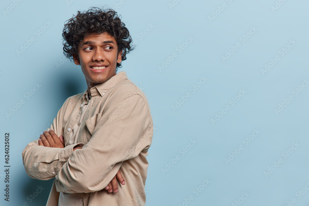Obraz premium Sideways shot of curly haired Hindu teenage man keeps arms folded looks behind smiles gladfully notices something pleasant isolated over blue background copy space for your advertising content