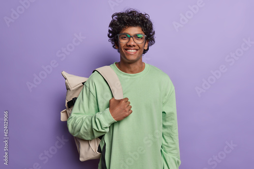 Photo of cheerful Hindu male backpacker or tourist smiles pleasantly leads active lifestyle and explores different places wears green jumper poses against purple background. Schoolboy daily life