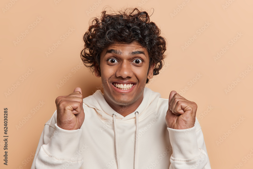Headshot of young Hindu man shows his clenched fists and radiant smile which symbolize triumph and accomplishment being in joyous state emphasizing victorious spirit wears casual hoodie poses indoor Headshot of young Hindu man shows his clenched fists and radiant smile which symbolize triumph and accomplishment being in joyous state emphasizing victorious spirit wears casual hoodie poses indoor