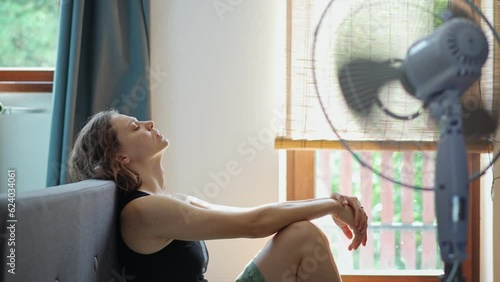 A young Caucasian woman tired from the summer heat sitting on the floor in front of a working electric fan.