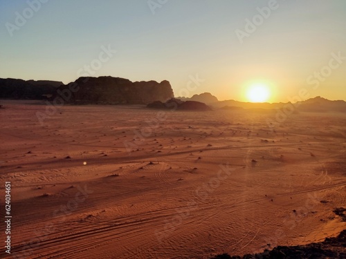Couchée du Soleil sur une dune rocheuse du désert de Wadi Rum, en Jordanie, plaine sableuse, promenade en jeep ou à dos de dromadaire, formation rocheuse rouge ou orange, avec trace de fumée jeeps