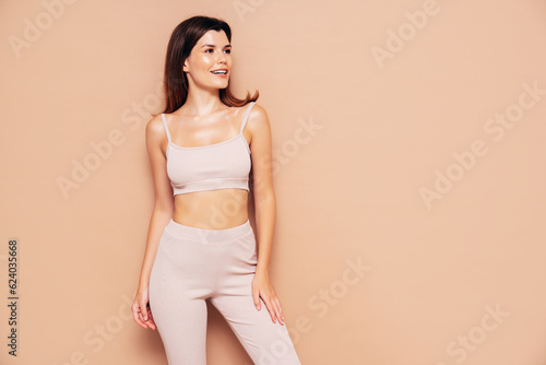 Young beautiful smiling female in trendy summer sport leggings and top clothes. Carefree woman posing near beige wall in studio. Positive model having fun indoors. Cheerful and happy. In sunglasses