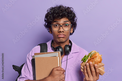 Discontent tired Hindu male student having rest in campus during lunch break carries notepads and sandwich dressed casually poses over purple background. Education and leisure concept. Back to school