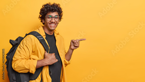 Sideways shot of cheerful curly haired male traveler with backpack pointing at copy space for nice travelling deal dressed casually isolated over yellow background. Travel lifestyle concept. © wayhome.studio 