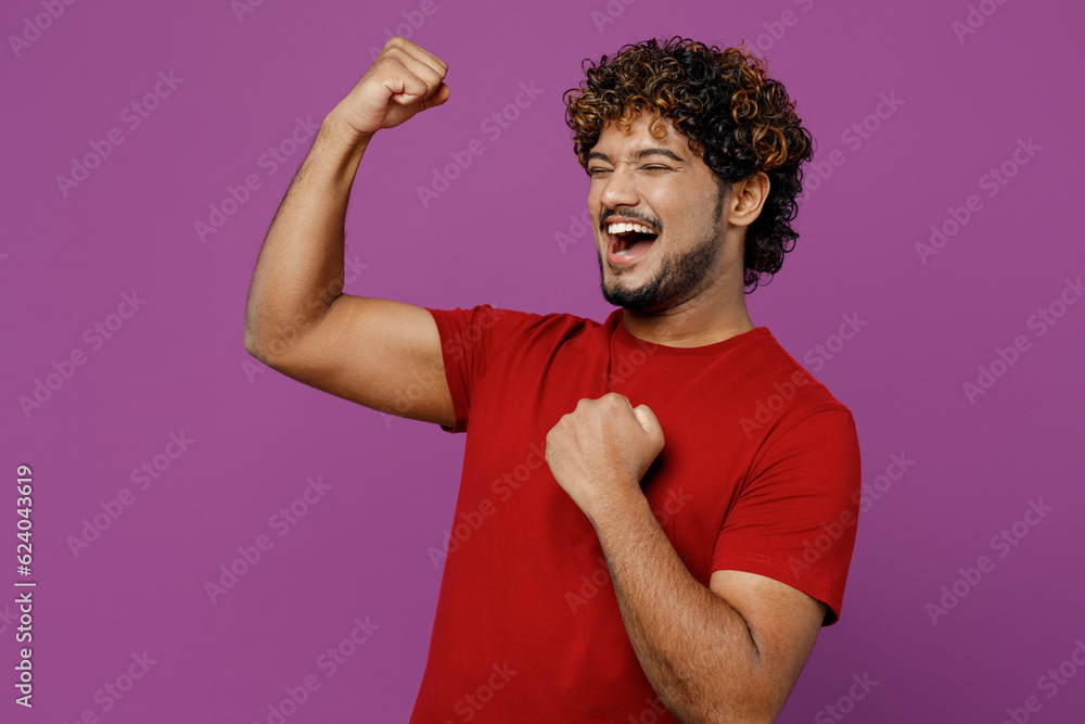 Side view young smiling happy Indian man wear red t-shirt casual clothes doing winner gesture celebrate clenching fists say yes isolated on plain purple background studio portrait. Lifestyle concept. Side view young smiling happy Indian man wear red t-shirt casual clothes doing winner gesture celebrate clenching fists say yes isolated on plain purple background studio portrait. Lifestyle concept.