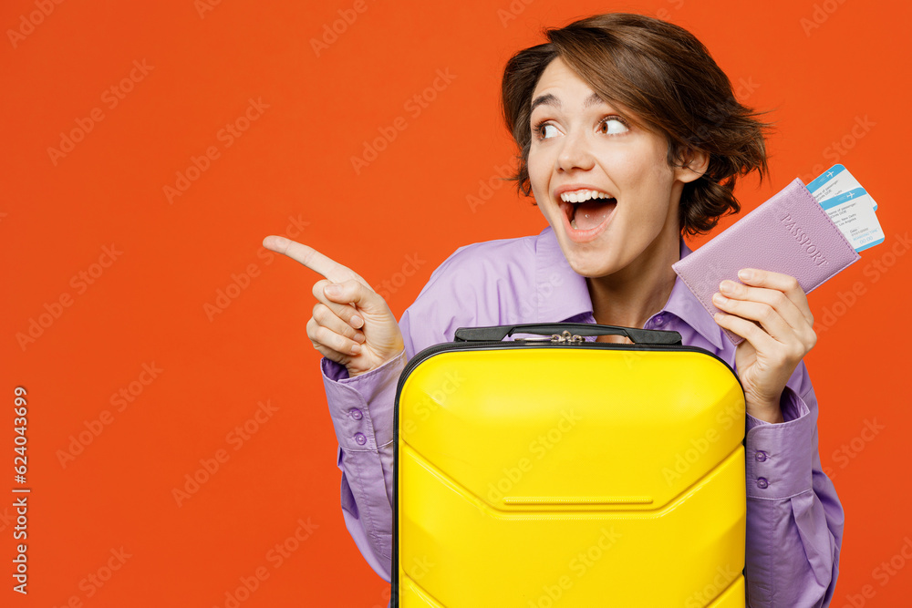 Traveler woman wear casual clothes hold suitcase passport ticket point aside isolated on plain orange background. Tourist travel abroad in free spare time rest getaway Air flight trip journey concept Traveler woman wear casual clothes hold suitcase passport ticket point aside isolated on plain orange background. Tourist travel abroad in free spare time rest getaway Air flight trip journey concept